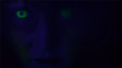 Dark photo of a dark blue femal face, almost completely in shadow, with one bright yellow iris visible.